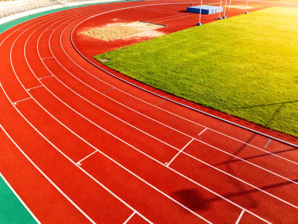 Widok z drona na profesjonalną bieżnię lekkoatletyczną z murawą i skoczniami, ukazujący standardy nowoczesnego obiektu sportowego.